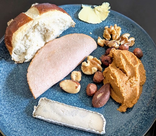 Ein blauer Teller mit einem halben Laugenbrötchen, veganem Kochschinken, Margarine, verschiedenen Nüssen, Tomatenhummus und einem Stück veganen Camembert auf Cashewbasis.