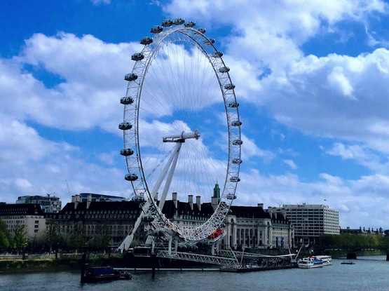 The London Eye or Millennium Wheel stands by the Thames River shore in London. Few large buildings are seen behind the wheel. Blue skies with white clouds above.  