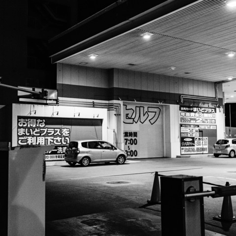 This black-and-white photograph captures the scene of a gas station at night. The station is well-lit, with bright lights illuminating the area and casting sharp shadows, which is characteristic of black-and-white photography.At the center of the image, there is a small car parked in front of a large, closed shutter door. Another car is parked on the right side of the photograph. The building features a flat roof that extends outward, providing a covered area for the vehicles. The architecture appears functional, with clean lines and a straightforward design.The ground surface is paved, and there are various signs and panels affixed to the building, though the specific content of the signs is not discernible in this description. There is a metal barrier arm in the foreground on the left side, likely used to control vehicle access to the area.The overall atmosphere of the scene is one of quiet and stillness, typical of a gas station at night, with no visible activity or movement.