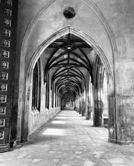Black and white photo of the cloisters at St John’s College. Repeating arches can be seen in a long line stretching out in front while sunlight is beaming in from the right side of the photo, casting interesting light patterns on the path. A large wooden door can be seen in the left side of the frame.
