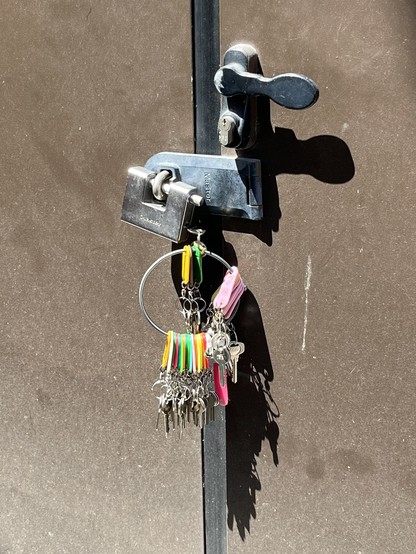 An industrial-looking faded brown metal door, with a silver-coloured handle and bar with a heavy duty padlock on it. There is a key in the padlock, attached to a large circular ring, with another maybe 30 or so keys attached, each with a brightly coloured plastic tag, in yellow, pale pink, bright pink, and neon green. The door is in direct bright sun, and the dark black shadows of the handle and key ring are surprisingly organic-looking, given the entirely man-made context. 