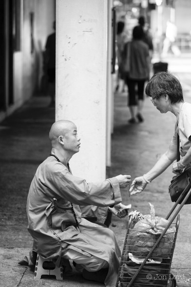 A Black And White Street Photo In Portrait Format. In The Lower Half Of The Image And Slightly Left Of Centre Sitting On A Small Stool Is A Young Buddhist Monk Handing Out Something To A Chinese Lady, Standing Along The Right Margin, Who Has Her Right Hand Stretched Out. In The Foreground And In The Right Lower Corner Is A Stacked Shopping Trolley. Behind the Monk And Lady Is A White Concrete Pillar With People Walking Away From The Camera Beyond And Blurred Out Of Focus. 

North Point, Hong Kong. 2014