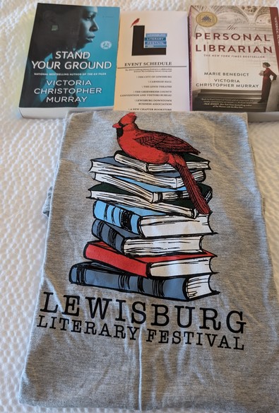Top left is a paperback copy of STAND YOUR GROUND by Victoria Christopher Murray, top center is the festival program, top right is a paperback copy of THE PERSONAL LIBRARIAN by Marie Benedict and Victoria Christopher Murray. Below the books is a gray T-shirt featuring a cardinal sitting on a stack of books, below the books are the words LEWISBURG LITERARY FESTIVAL.