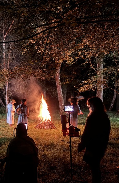 Nighttime. A log fire in a small clearing in the woods. Several figures wearing crowns of thorns, twigs and flora, dressed in white dance around. Camera crew and director look on. 