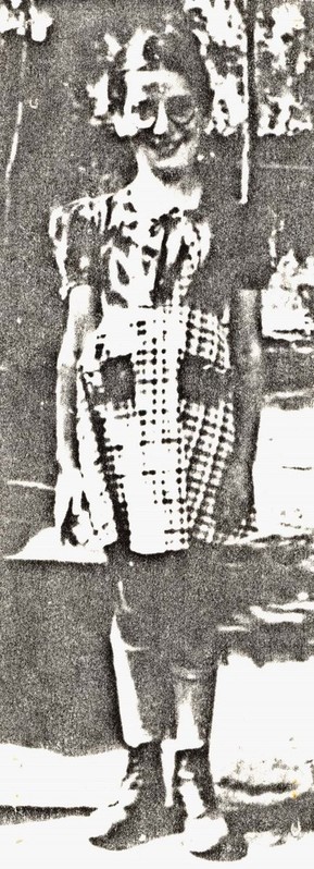 A black and white vintage photo of a girl standing outdoors in a patterned dress, smiling at the camera. The background is indistinct, with lighter areas suggesting foliage or trees.
