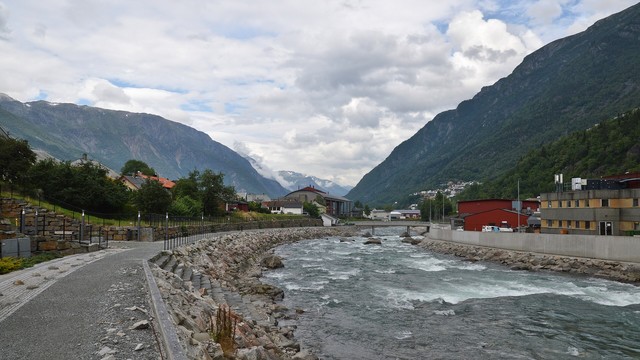 A photo of a river going through a small town. There is a path to the side of the river and the mountainous sides of fjords in the distance. The sky is filled with white clouds.
