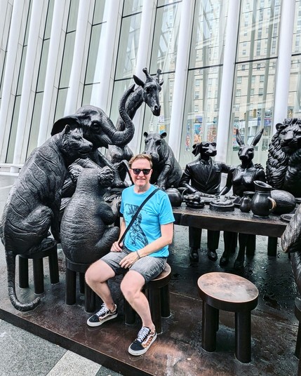 Me (wearing a turquoise Madonna T-shirt and black denim shorts) sitting in front of a sculpture of several animals around a table, including a koala, elephant, giraffe, hippo and lion