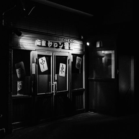 In the black-and-white photograph, a building with a wooden facade is depicted. The structure features a pair of glass double doors at its center, with each door framed by wooden panels. The glass sections of the doors have opaque coverings, likely to obscure the view of the interior. On each of these coverings, there are visible shapes resembling cards or placards, each with symbols or characters inscribed on them. There are also smaller posters or signs partially visible around the doors, affixed to the glass or wooden surfaces.The setting appears to be dark, with only a few sources of light illuminating the scene. One light source is positioned above the doors, casting shadows and highlighting the textures of the wood and glass. The light creates a contrast between the illuminated areas and the darker surroundings, emphasizing the structure’s details and the objects attached to it. There are no people visible in the photograph, and the surrounding environment is not detailed, keeping the focus on the entrance of the building.