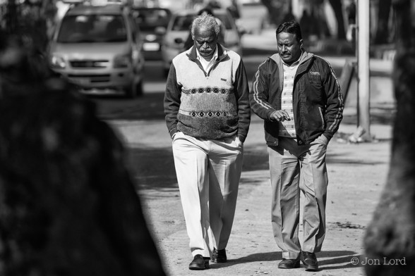 A Black And White Street Photo In Landscape Format. In The Centre Of The Image In An Indian Man Walking Along The Pavement, Looking Down As He Walks. He Has Grey Hair And A Grey Moustache. He Is Wearing A Patterned Sweater And Has Light Coloured Trousers. To The Right Is Another Indian Man, Looking Slightly Towards The First Man With One Arm Slightly Outstretched As If Making A Point. He Has Jet Black Hair, Parted In The Centre And Has A Small Dark Moustache. He Is Wearing A Dark Casual Jacket. On The Left And Right Margins Are The Dark Outlines Of Trees On The Roadside. In The Background Is Traffic, Blurred Out Of Focus And Coming Towards The Camera. The Location Is Saket, New Delhi. The Image Was Shot In January (It Was Cold) 2013.