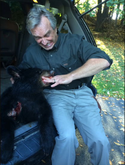 RFK Jr. sitting in the open back of a minivan or SUV, with his hand in the mouth of the dead bear cub, pretending the dead bear cub is biting him and grimacing