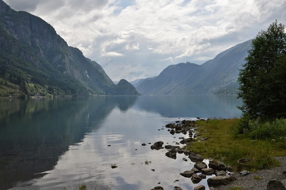 A photo of a lake surrounded by mountains. Some of the near shore is visible on the right side of the image and a tree growing out of it. The sky is filled with white and grey clouds.