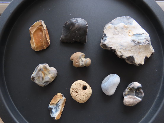 Photo of a collection of rocks on a black matt round table. Mostly black and brown flints. One piece with a hole right through may be remnants of a house brick.