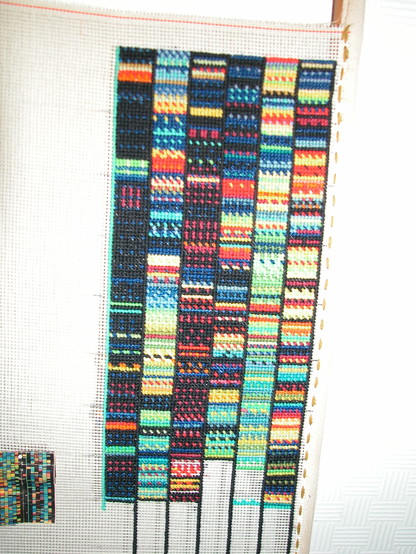 Photo of a work in progress, tapestry on a frame. Part of the human genome with the small image bottom left cut from a magazine. Multiple short bands of colours in rows separated by rows of black stitches.