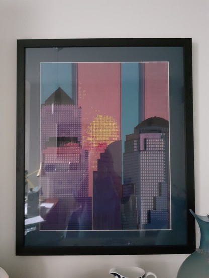 Photo of a large framed tapestry of the Twin Towers New York with the sun reflected in one of them and an unfinished roof of a building in the middle left. Done with tapestry cotton.