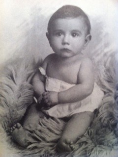 Black and white vintage photograph of a baby boy sitting on a fluffy fabric, wearing a light-colored outfit and looking directly at the camera.
