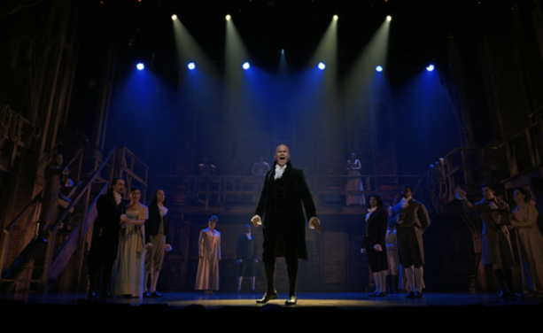 Full stage view of Hamilton (the musical). Christopher Jackson is center stage and forward singing One Last Time. In the background, most other cast members are scattered around behind in semidarkness. 