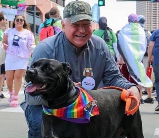 A smiling man wearing a gray jacket and cap kneels beside a black dog adorned with a rainbow bandana. They are surrounded by people in colorful attire, indicating a festive or pride event.