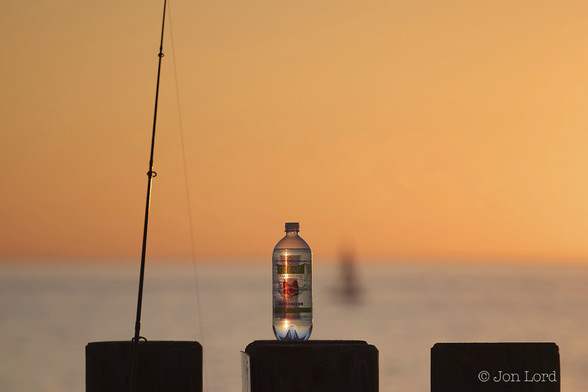 A Colour Abstract Photo In Landscape Format. In The Foreground At The Base Of The Image Are The Tops Of Three Wooden Posts. The Post On The Left Has A Fishing Rod Resting On It And Is Reaching Out Of View At The Top. The Line Is Visible Hanging Downwards. On The Middle Post, Which Is Also In The Centre Of The Photo, Is A Bottle Of Mineral Water, The Bottle Is Transparent And The Golden Glow Of The Setting Sun Can Be Seen Through It. Slightly To The Bottles's Left Is The Silhouette Of A Distant Sailing Boat, Blurred Out Of Focus. On The Third Post: Nothing. About One Third Up From The Bottom Is The Horizon Where The Pacific Ocean Meets The Sky. Above Is A Clear And Cloudless Sky, Golden In The Setting Californian Sun.

Redondo Beach, California. 2013