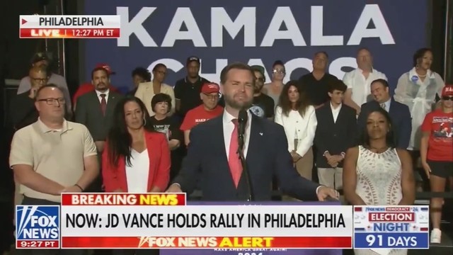 JD Vance at a campaign rally standing in front of a banner that reads “Kamala Chaos” except the crowd behind him is covering the second word.