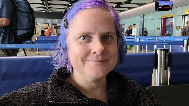 Woman with black jumper and purple hair sitting in an airport.