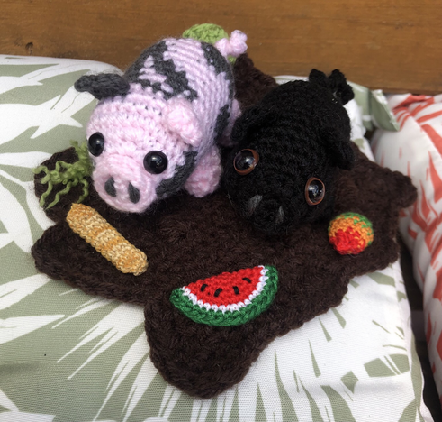 Photo shows two small crochet pigs, one pink and grey, the other is smaller and black. They have a crochet mud splat with crochet food - watermelon, baguette, cabbage, mango and grass tufts.