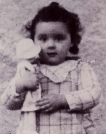 A black and white photo of a young girl holding a doll, wearing a plaid dress and looking directly at the camera. The photo is slightly blurred and has a vintage quality.
