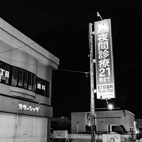 This black and white photograph captures a nighttime urban scene in Japan. The image is dominated by a large illuminated sign written in Japanese characters, mounted on a tall pole. The sign appears to be advertising a medical or dental service, with numbers visible at the bottom. To the left of the sign is a multi-story building with a curved facade. The building's windows are dark, suggesting it is closed. Below the windows, there is a store sign in katakana characters. The overall atmosphere is quiet and still, typical of a late-night street view in a Japanese town or city. The contrast between the brightly lit sign and the dark surroundings creates a stark visual effect. In the foreground, part of a parked vehicle is visible. The grainy texture and high contrast of the image contribute to its moody, film noir-like quality, emphasizing the emptiness and silence of the night.