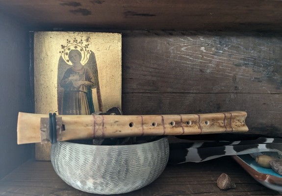 A flute made from a deer femur in a wooden niche placed atop a silver bowl with a shiny gold angel icon in the background, an acorn, and a couple of bird feathers, one from a crow and one from a coopers hawk.