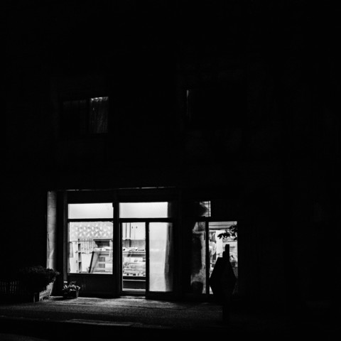 This black and white photograph captures a nighttime scene of a small storefront or shop. The image is dominated by darkness, with the main focus being a brightly illuminated window. The window appears to be part of a small building or structure, likely a convenience store or similar retail establishment. Through the window, shelves and products can be faintly seen, suggesting it is indeed a shop interior. The storefront has a glass door, which is closed, and the entrance area is lit, creating a stark contrast with the surrounding darkness. A small section of pavement is visible in front of the store, and there appears to be some kind of small object, possibly a plant pot, near the entrance. Above the main window, there is another smaller, dimly lit window visible in the upper left corner of the image. The overall composition creates a sense of isolation and quietness in what seems to be a late-night urban setting.