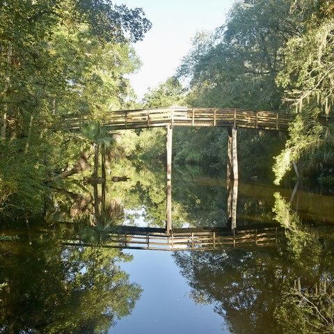 Wooden foot bridge on high posts arching across river with trees and sky, all reflected below in river