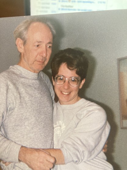 my father and I standing up holding one another. His gaze is straight ahead. I am looking at the camera smiling, happy to be with him. We are both wearing gray sweatshirts.