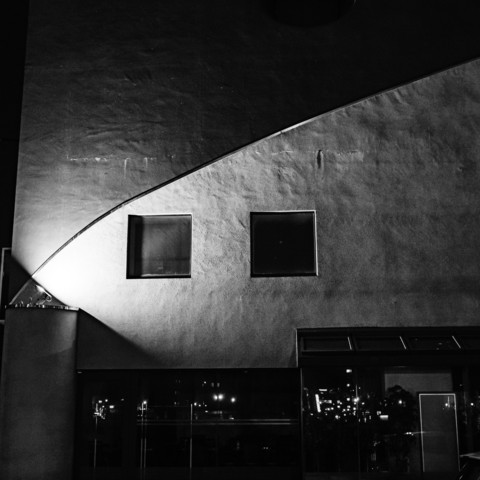 This black and white photograph captures a striking architectural scene. The image is dominated by a large, textured wall with two square windows. The wall's surface appears rough and uneven, possibly concrete or stucco. A diagonal line cuts across the upper left corner of the frame, creating a dramatic contrast in light and shadow. The two windows are perfectly aligned and appear dark against the lighter wall. Below the wall, there's a large glass panel or window that reflects city lights, suggesting this is an urban nighttime scene. The reflection shows scattered points of light, likely from street lamps or buildings in the distance. The composition is geometric and minimalist, emphasizing the interplay of light, shadow, and architectural elements. The high contrast and grainy texture of the image enhance its moody, atmospheric quality.