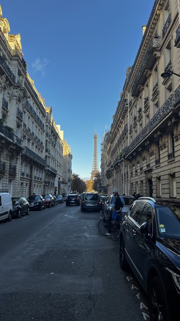 A view of a street in Paris, featuring classic buildings lining both sides, with the Eiffel Tower visible in the distance under a clear blue sky. Several parked cars are along the road, and there is a cyclist and a pedestrian in the scene.