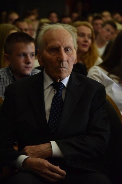 Kazimierz Piechowski seated in an auditorium, wearing a suit and tie, surrounded by audience members.