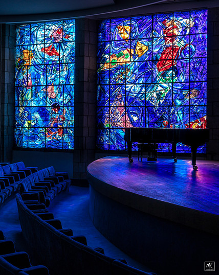 Color photo of an auditorium with curved rows of seats before a raised stage with a piano it.  The wall beyond has two huge colorful stained glass windows designed by Marc Chagall and the colored light coming through them is dimly illuminating the interior of the auditorium. 