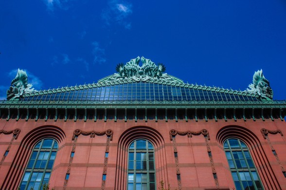 Chicago Public Library, Harold Washington Branch. The red brick building is several stories high. There are 3 towering windows that end in a rounded arch at the top. Stonework that resembles fabric bunting is spaced between the windows. The building is crowned with a green metal structure, that may be painted or verdigris. The green metal roof contains a triangular section made of glass windows attended in small squares. At the roof peak and at either end of the slanted roof, there are metal sculptures that look like very sharp Scottish thistles. There is a bright blue sky above the building and that is reflected in the library windows.