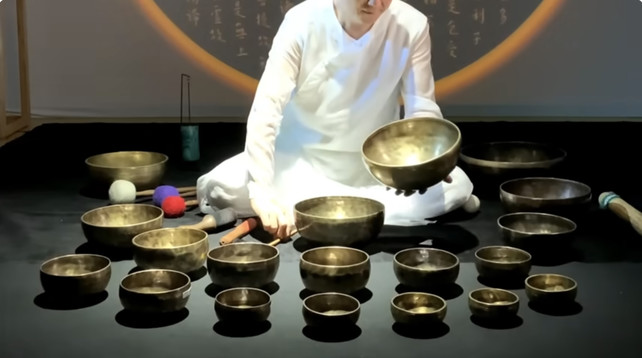 This photo is a screenshot from a YouTube video. It shows a man wearing white seated on the floor. In front of him is a large collection of Tibetan singing bowls, and the man plays the bowls musically to produce an otherworldly sound, one that is half science fiction soundtrack and half heavenly ceremony. 