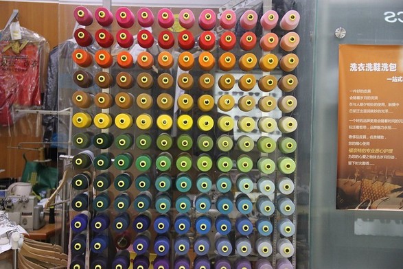 A cheerful rectangular display of differently colored spools of thread in front of a dry cleaners and tailor shop represents all the hues of the rainbow, starting from pink, red, and orange at the top; to brown, yellow, and green in the middle; and shading into aqua, blue, indigo, purple, and lavender at the bottom.