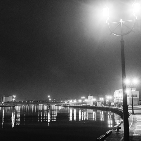 This black and white nighttime photograph captures a waterfront scene with a curved promenade or harbor walkway. Street lamps with distinctive half-circle designs line the path, casting bright light that reflects brilliantly on the calm water's surface. The reflections create long, vertical streaks of light across the water, forming a mirror-like pattern. Several buildings are visible along the waterfront, including what appear to be commercial structures and a taller apartment building in the distance. The curvature of the walkway follows the natural contour of the waterfront, with a protective barrier or railing separating pedestrians from the water. The composition emphasizes the contrast between the dark night sky and the illuminated elements, with the street lamps prominently featured in the foreground. The overall atmosphere is serene and urban, with the artificial lighting creating a dramatic interplay of light and shadow across the scene.