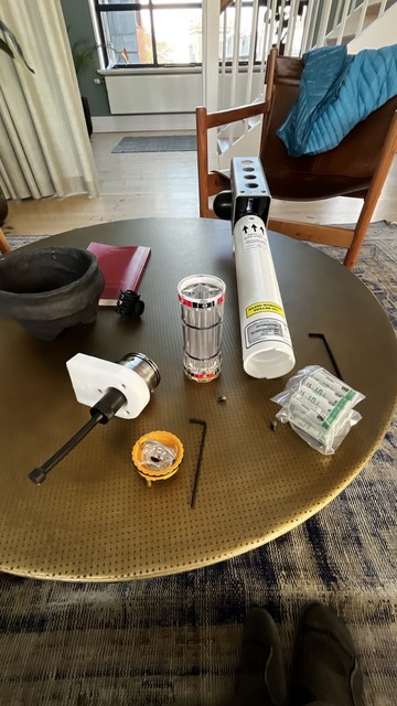 A circular table displays various tools and components, including a round container, two small transparent tubes, a cylindrical white tube, several hex keys, and a stack of batteries. A red notebook and a chair with a blue jacket are visible in the background.