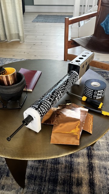 A tabletop scene featuring a device with a black and white patterned tube, a metallic component, a roll of PVC tape, a screwdriver, and several sheets of shiny copper foil. Nearby, there is a black bowl, a books, and a pair of glasses. 