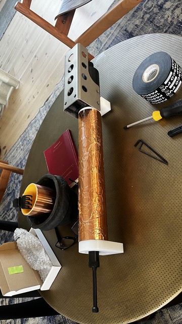 An assembly of mechanical components on a round table. Items include a metal part with holes as part of a large copper-colored tube, a black container, a roll of copper tape, a screwdriver, PVC tape, and a small box. The background features wooden flooring.