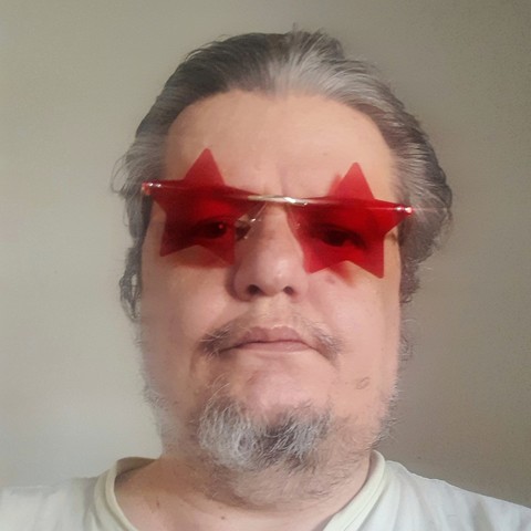 Selfie de um homem idoso feio. Branco, rosto redondo, cabelos na maior parte brancos, testa enorme, barba rala na maior parte grisalha, bigodinho fino ridículo, usando óculos com lentes em forma de estrelas vermelhas e camiseta desbeiçada verde