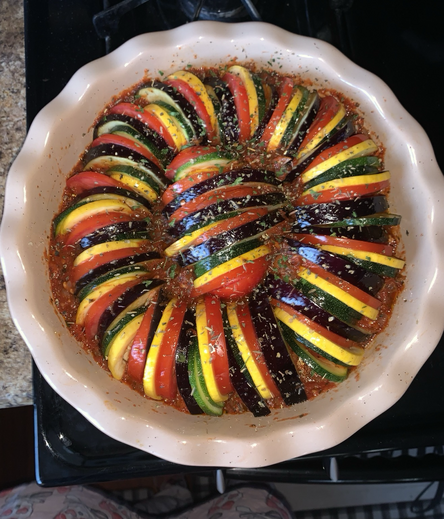 A neutral colored ceramic pie dish. Inside is a ring of sliced vegetables, nestled inside of a tomato based sauce, sprinkled with herbs. The sliced vegetables are arranged in alternating order of eggplant, purple, Roma tomato, red, squash, yellow, and zucchini, green. This image is of the dish prior to cooking.