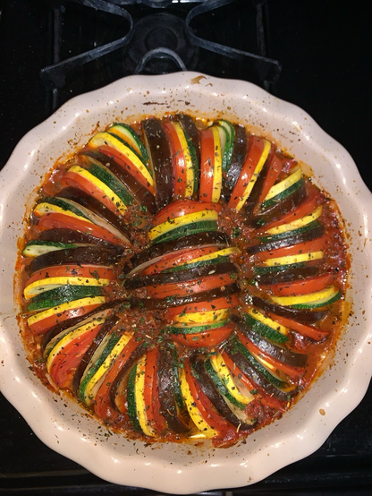 A neutral colored ceramic pie dish. Inside is a ring of sliced vegetables, nestled inside of a tomato based sauce, sprinkled with herbs. The sliced vegetables are arranged in alternating order of eggplant, purple, Roma tomato, red, squash, yellow, and zucchini, green. This image is of the dish after cooking, but looks almost the same. The visual difference is the colors of the vegetables are slightly more vivid & the sprinkled herbs are dark & wilted. 