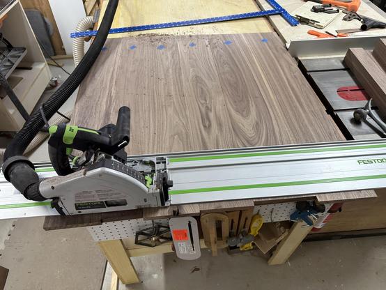 A woodworking setup featuring a circular saw positioned on a guide rail, with a large sheet of wood and various tools and materials arranged on a workbench. The workspace includes a vacuum attachment for dust collection and multiple clamps visible nearby.