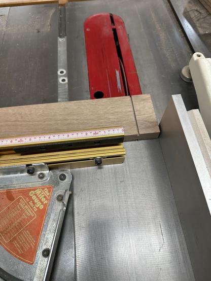 A close-up of a table saw workspace, showing a piece of wood being measured against a ruler. The saw blade and a metal miter gauge are visible, indicating woodworking in progress.