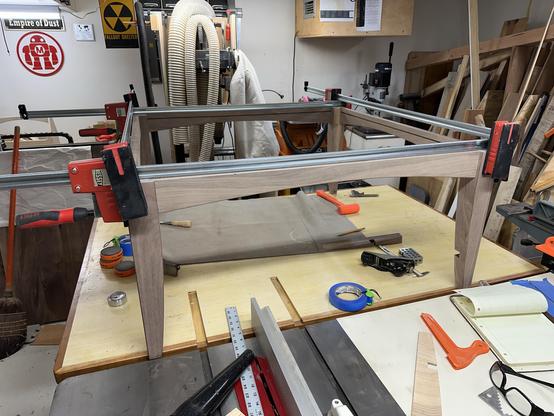 A woodworking workspace featuring a partially assembled wooden frame clamped to a table. Surrounding tools and materials include clamps, a roll of tape, a square, and various wood pieces. Background elements include machinery and a wall sign.