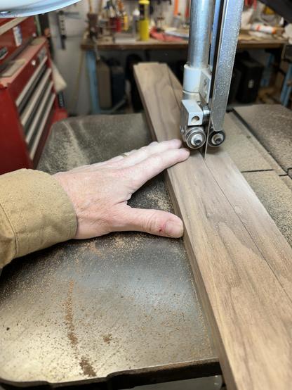 A hand is positioned on a wooden plank being cut on a bandsaw in a workshop. Sawdust is visible on the surface, and tools are in the blurred background.