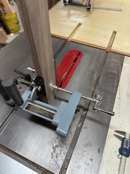 A woodworking table saw setup featuring a vertical piece of wood being secured with a clamp. The saw blade and a guide rail are visible, along with a caliper and other tools on the table.